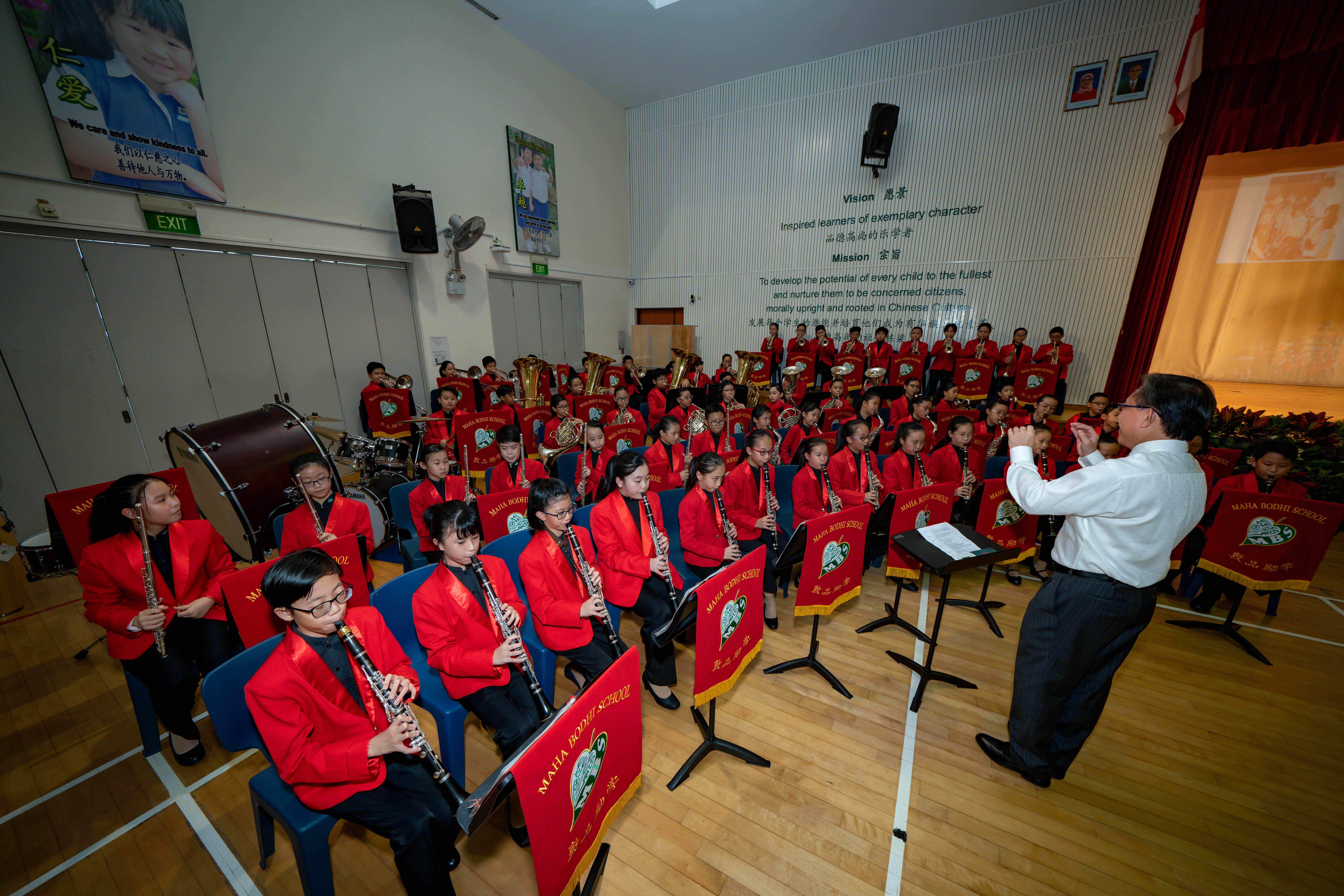 Students in red blazers play wind instruments in a school band, led by a conductor at a music stand.